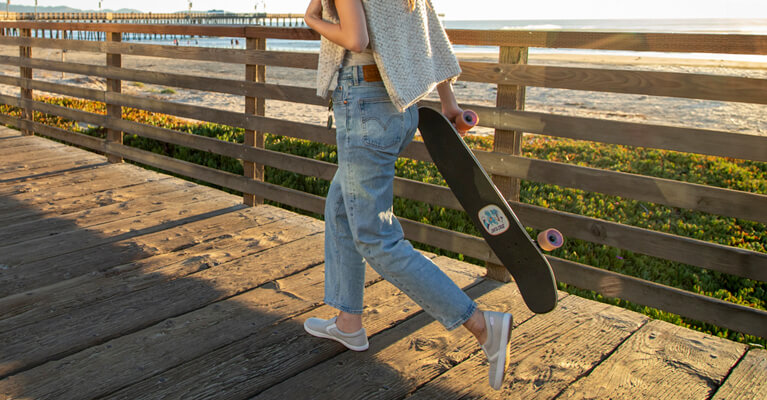 A person walking down the boardwalk in their Dillon Canvas Slip Ons holding a skateboard.