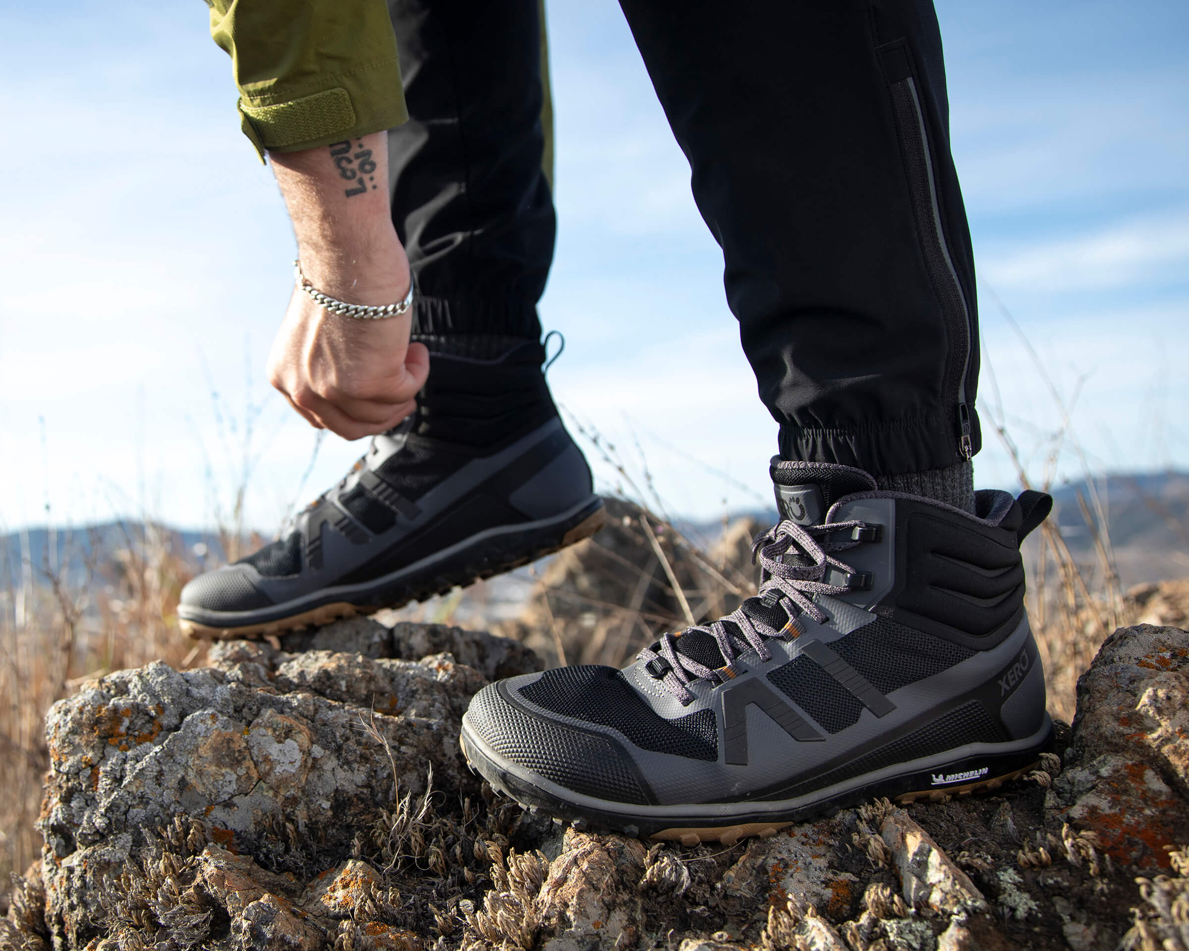 Ein Mann passt die Schnürsenkel seiner Scrambler Mid II-Stiefel an, während er draußen auf Felsen steht