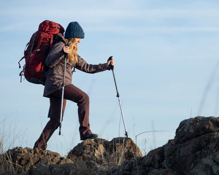 Eine Frau in Scrambler Mid II-Wanderstiefeln wandert an einem klaren, kühlen Tag mit einem großen Rucksack und Trekkingstöcken über Felsen.