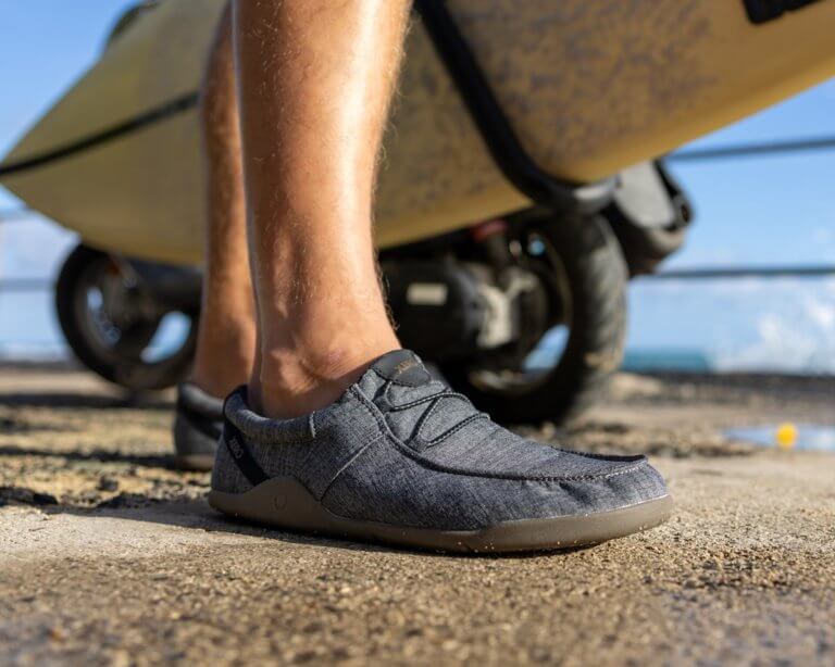 Lower leg view of a man wearing Kona beach-style slip-on shoes standing in front of a surf board cart