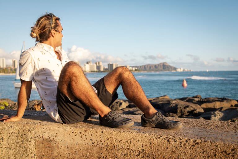 A man wearing Kona beach-style slip-on shoes sitting on concrete wall looking at the ocean