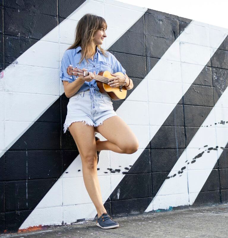 Žena opřená o zeď hraje na ukulele a má na sobě plážové boty Kona.