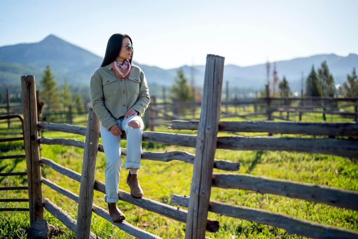 A woman casually sitting on a wooden fence wearing Ridgeway Chelsea boots with mountains and trees in the background