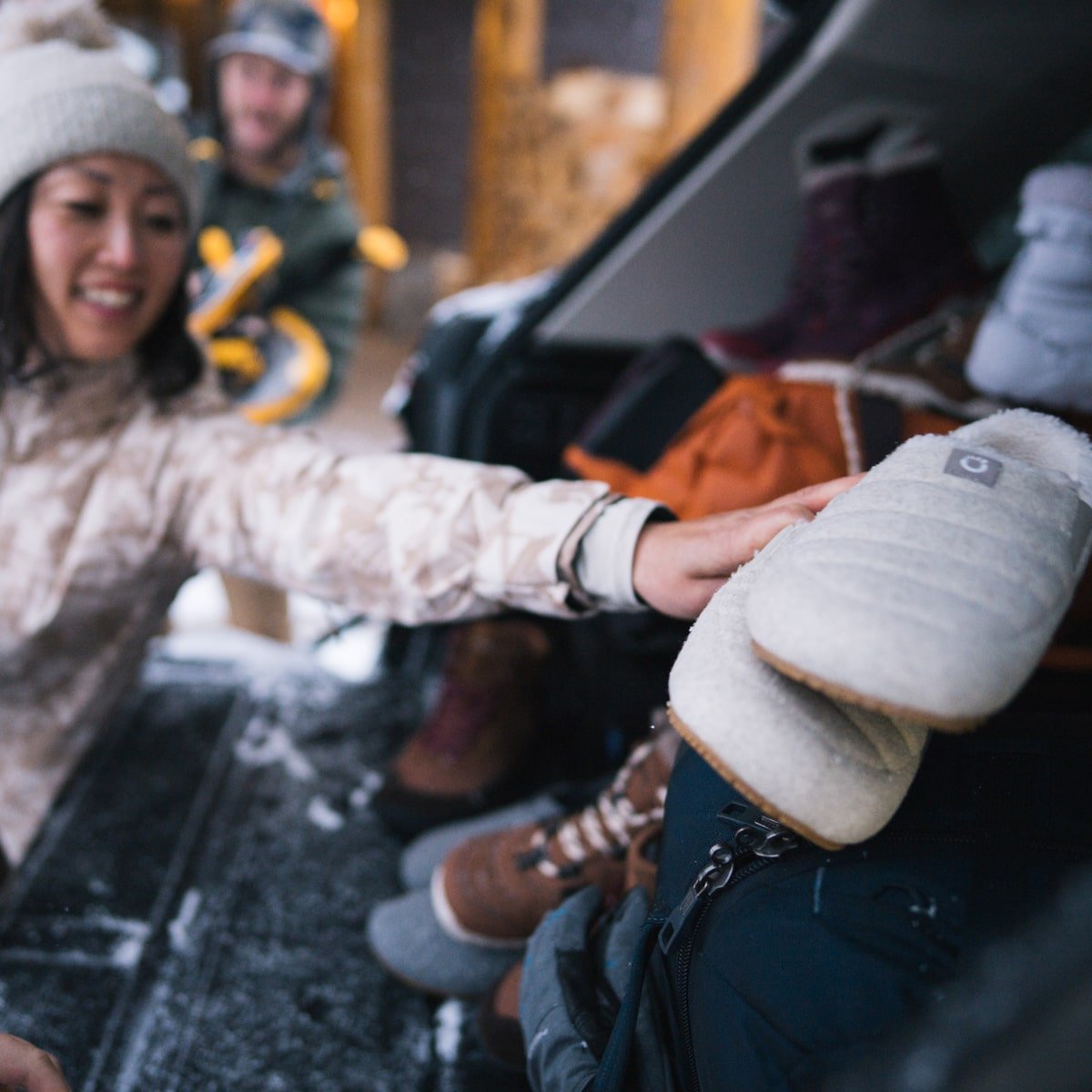 Woman reaching into a car trunk outdoors, holding a pair of Pagosa Cozy marled cream slippers in snowy setting