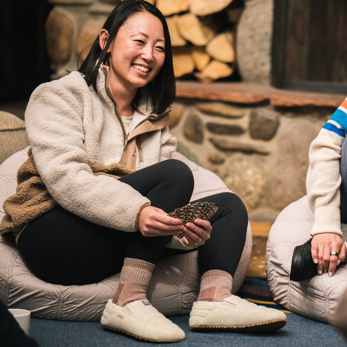 Woman smiling and playing cards indoors, wearing Pagosa Cozy marled cream slippers with pink socks, sitting by a fireplace
